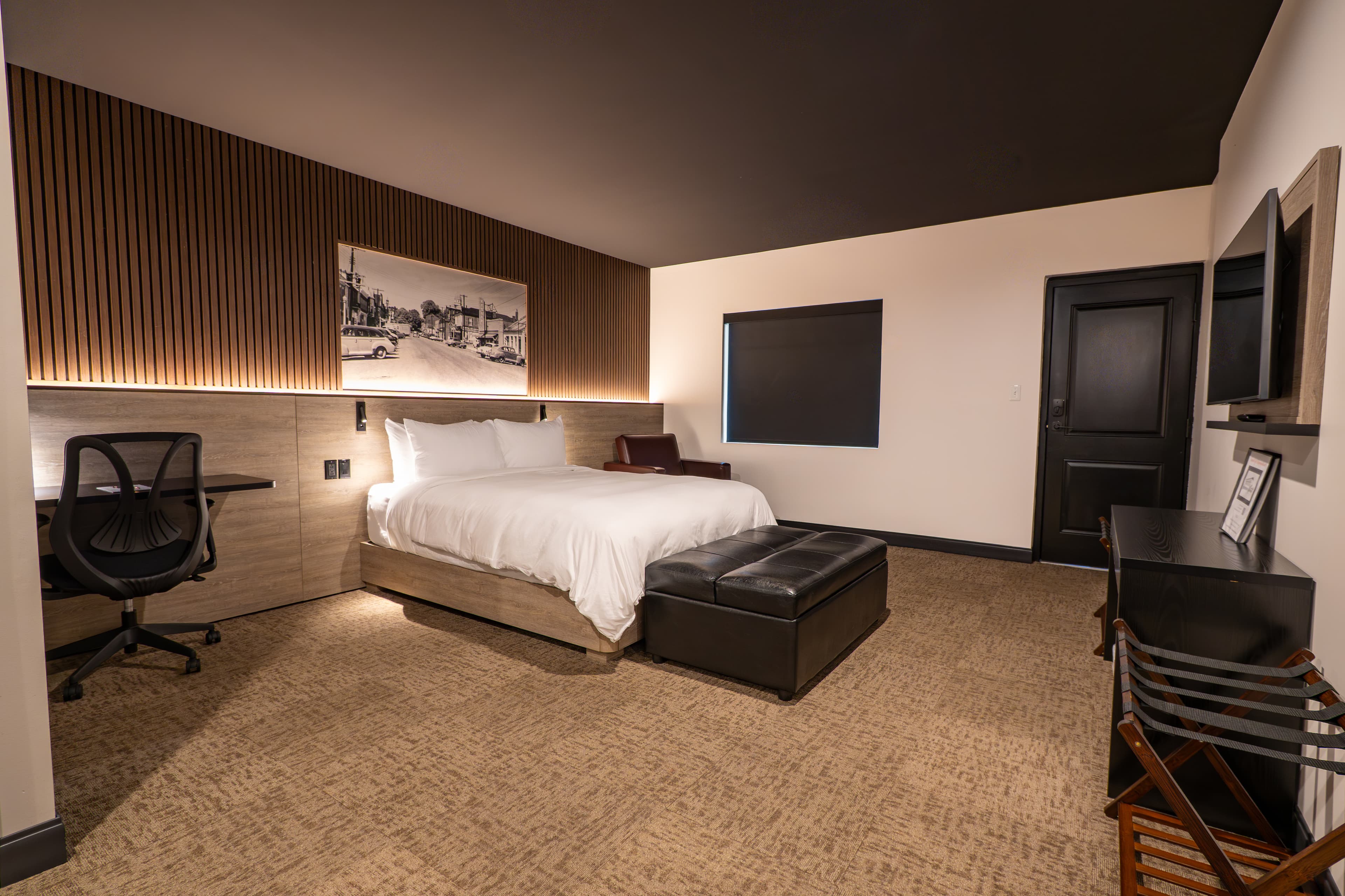 A modern hotel room featuring a queen bed, desk, and seating area with warm lighting and minimalist decor.
