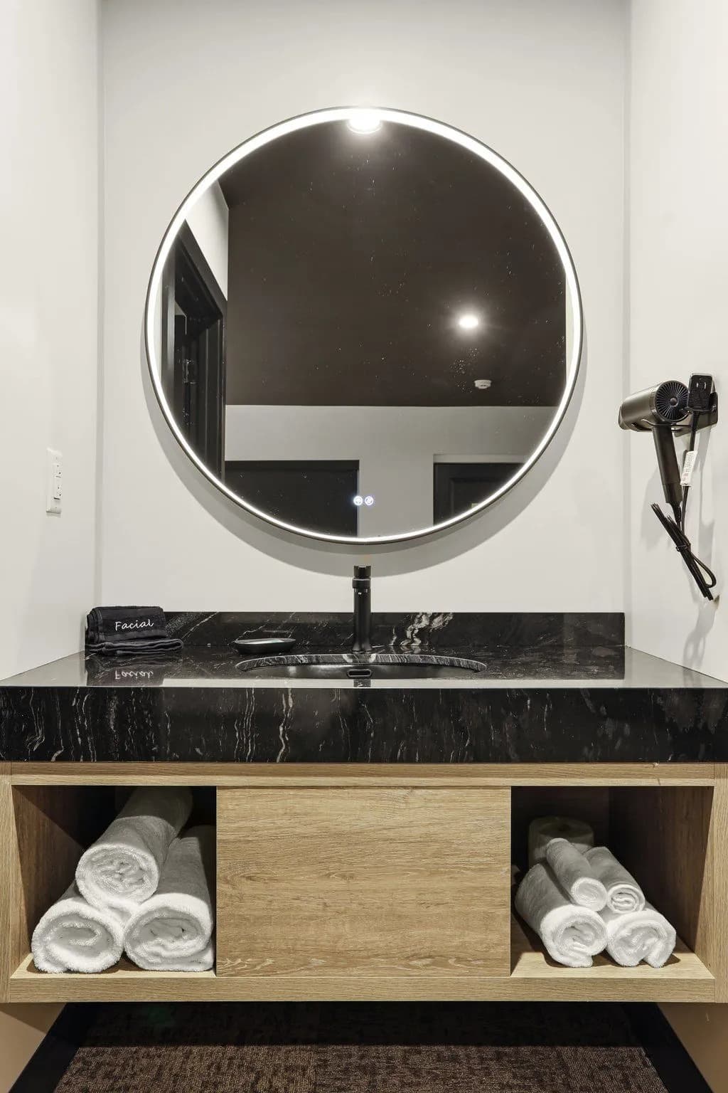 A modern bathroom sink with a circular mirror, hair dryer, and neatly stacked white towels in wooden shelves below.
