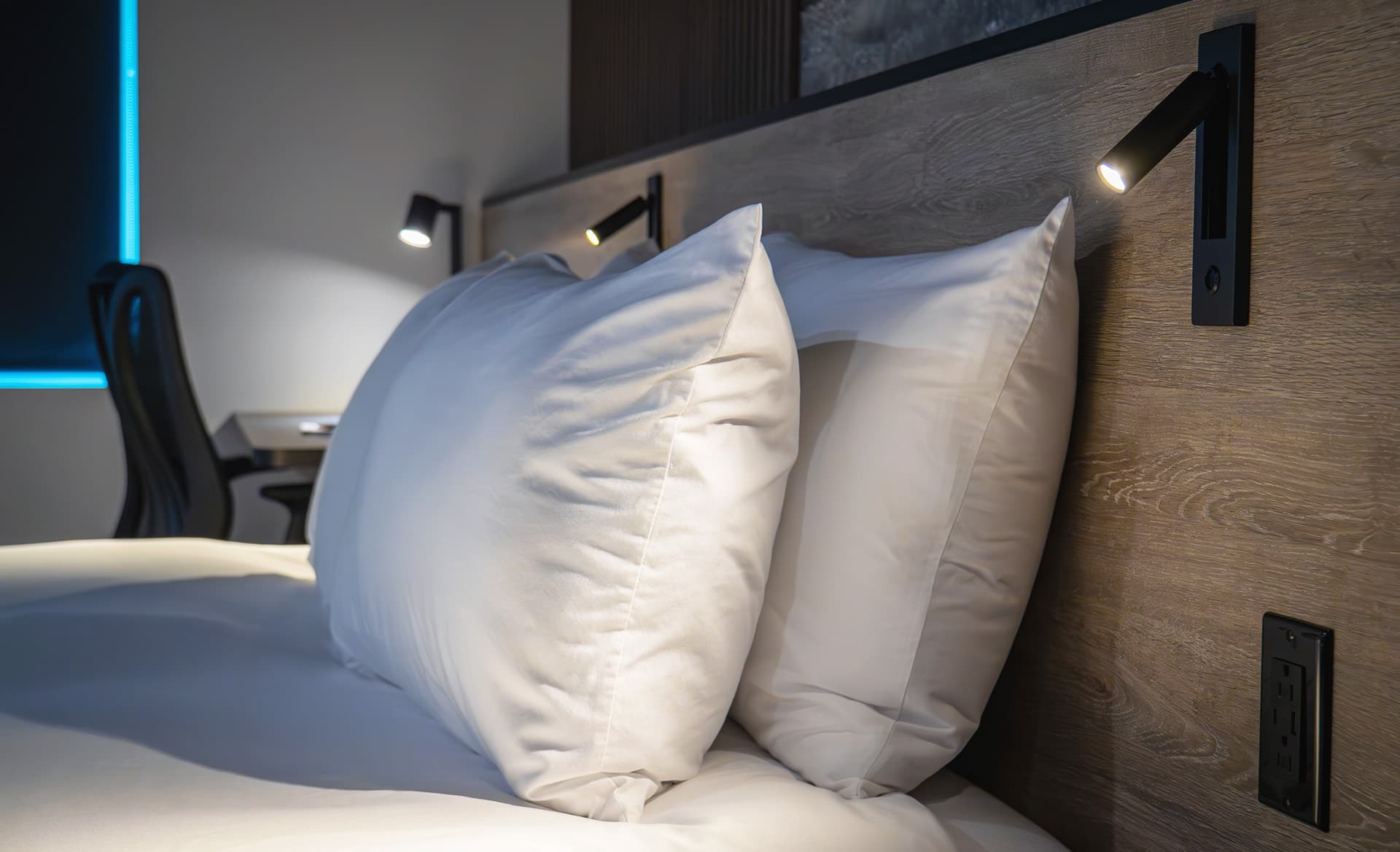 A neatly made bed with two large white pillows against a wooden headboard and wall-mounted reading lights.