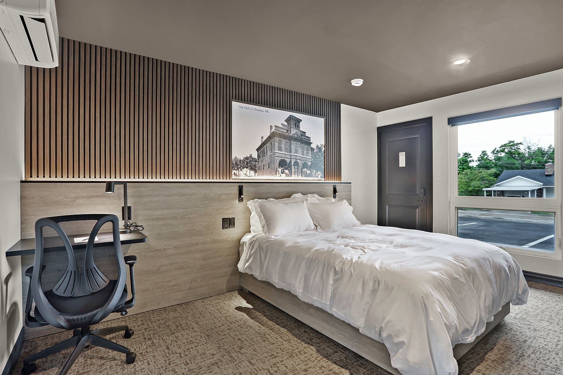 A modern bedroom featuring a white bed, a desk chair, wooden paneling, and a large window.