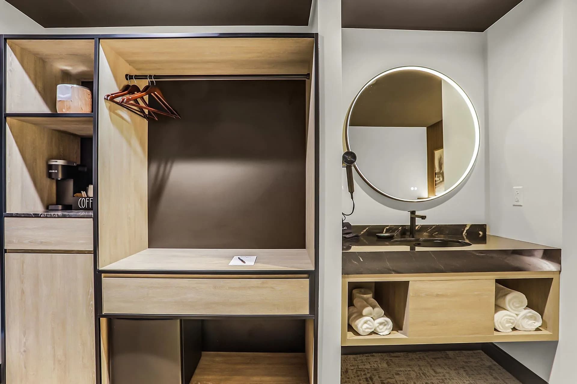 A modern hotel room closet and vanity area featuring a round mirror, hanging clothes, and neatly stacked towels.