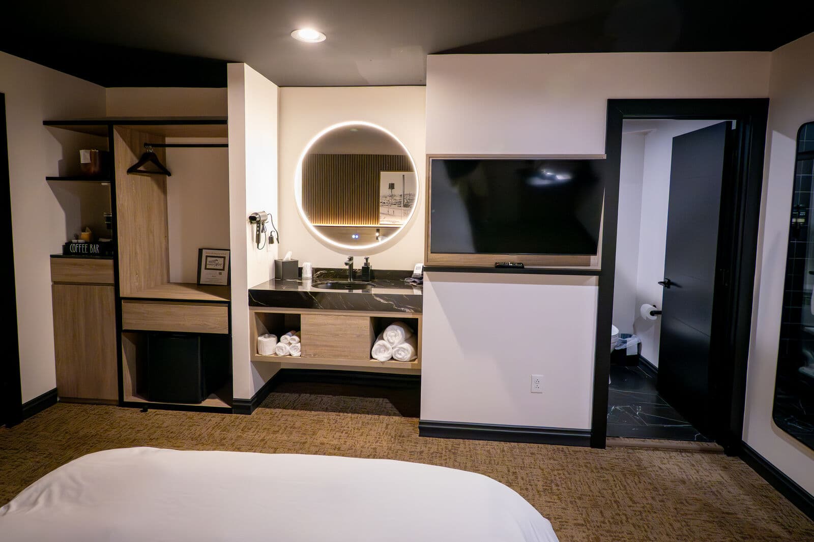 A modern hotel room featuring a round mirror, television, sink area, and an adjoining bathroom.