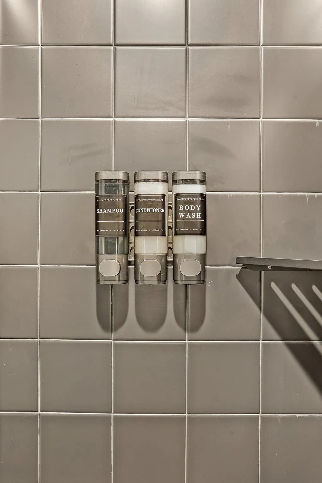 Three neatly arranged dispenser bottles labeled "Shampoo," "Conditioner," and "Body Wash" attached to a gray tiled wall.