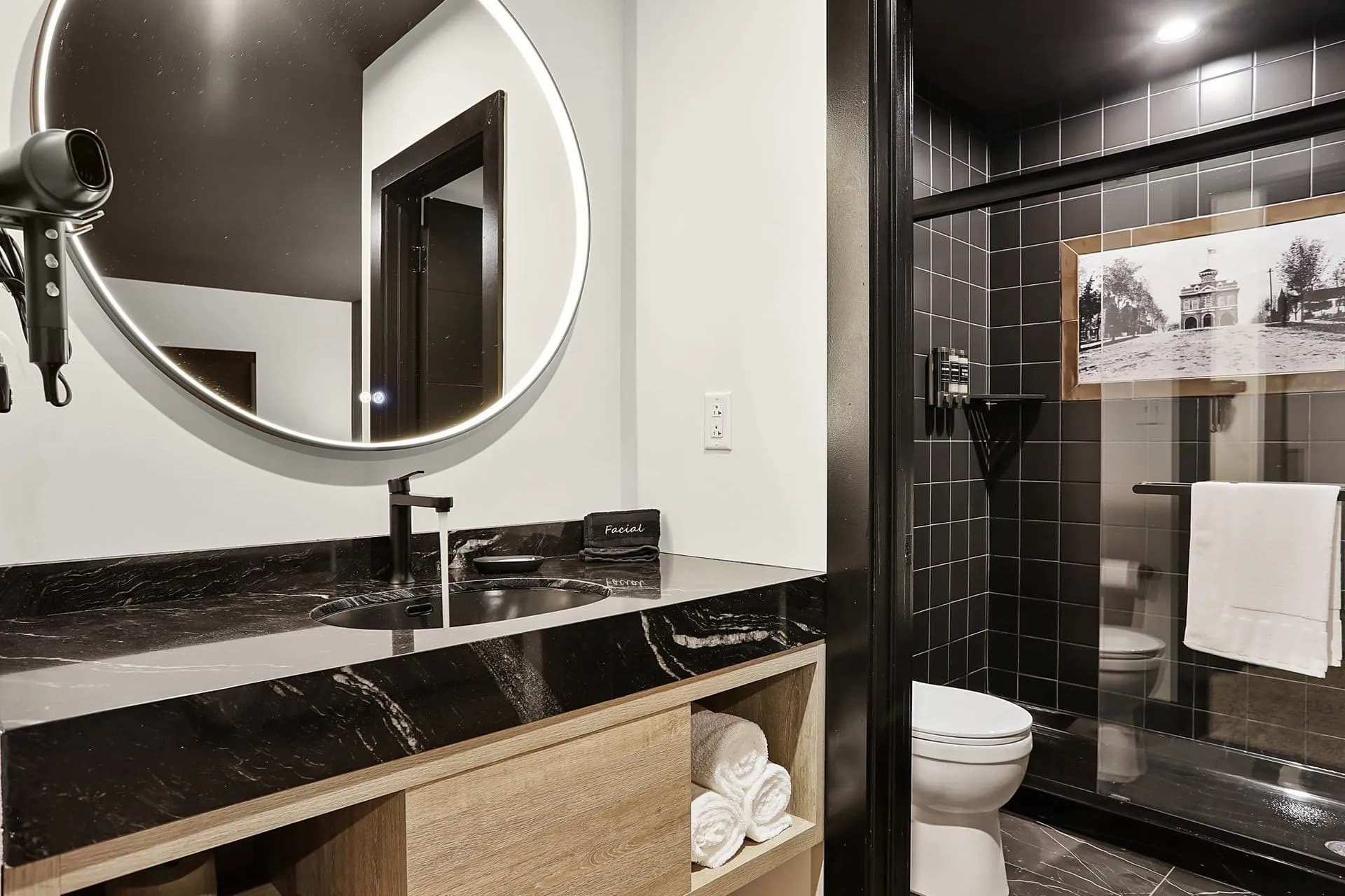 Modern bathroom featuring a circular mirror, dark tile shower, wooden vanity, and neatly stacked towels.