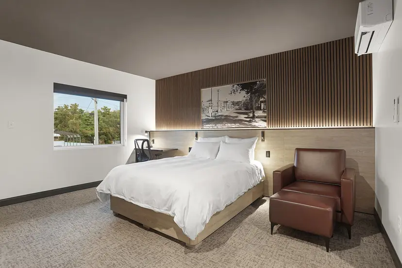 A modern hotel room featuring a queen bed, armchair, desk, and large window with a view.
