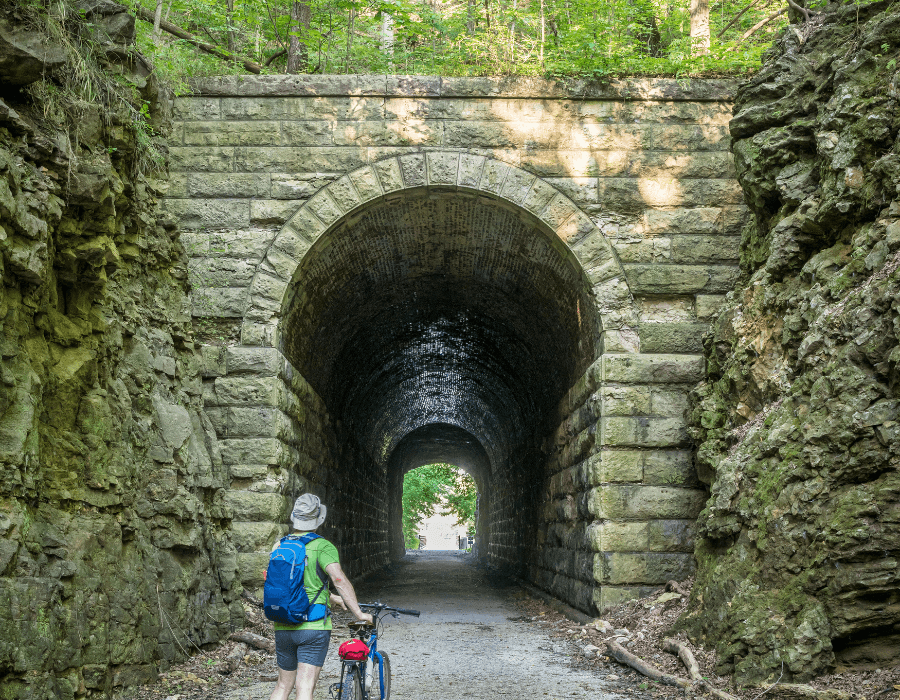 A cyclist in a hat stands in front of a stone tunnel surrounded by lush greenery.
