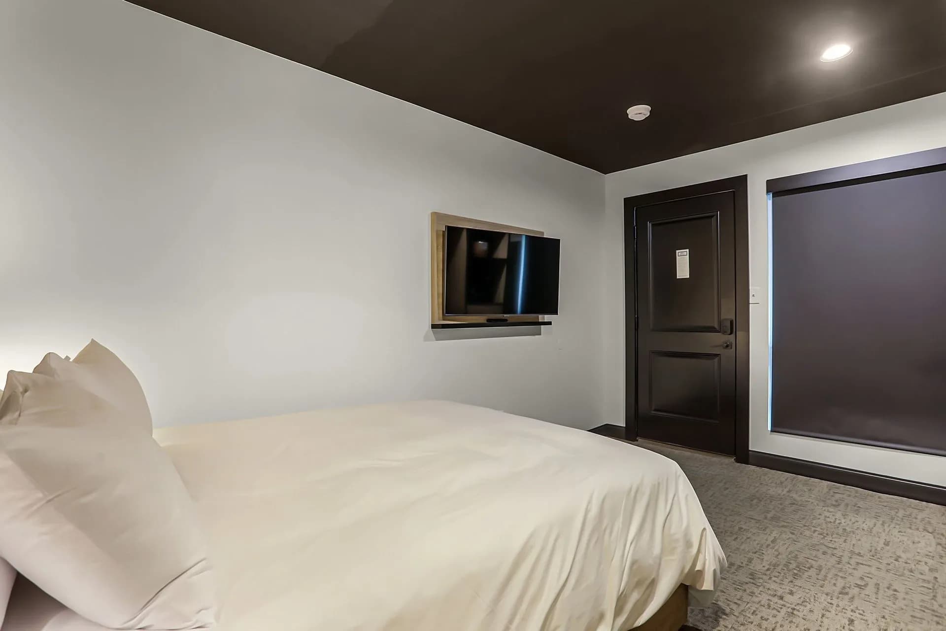 A minimalist hotel room featuring a bed, a wall-mounted TV, and a door with a black-out window shade.
