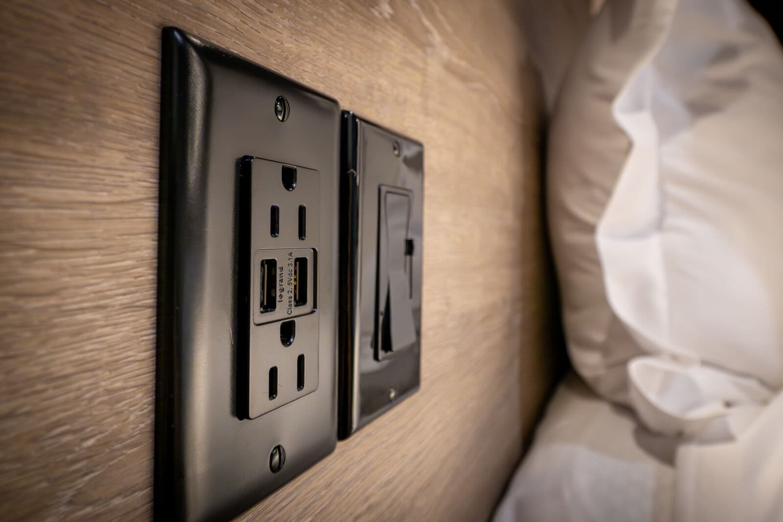 A close-up of a black electrical outlet featuring USB ports, mounted on a wooden wall beside a bed.