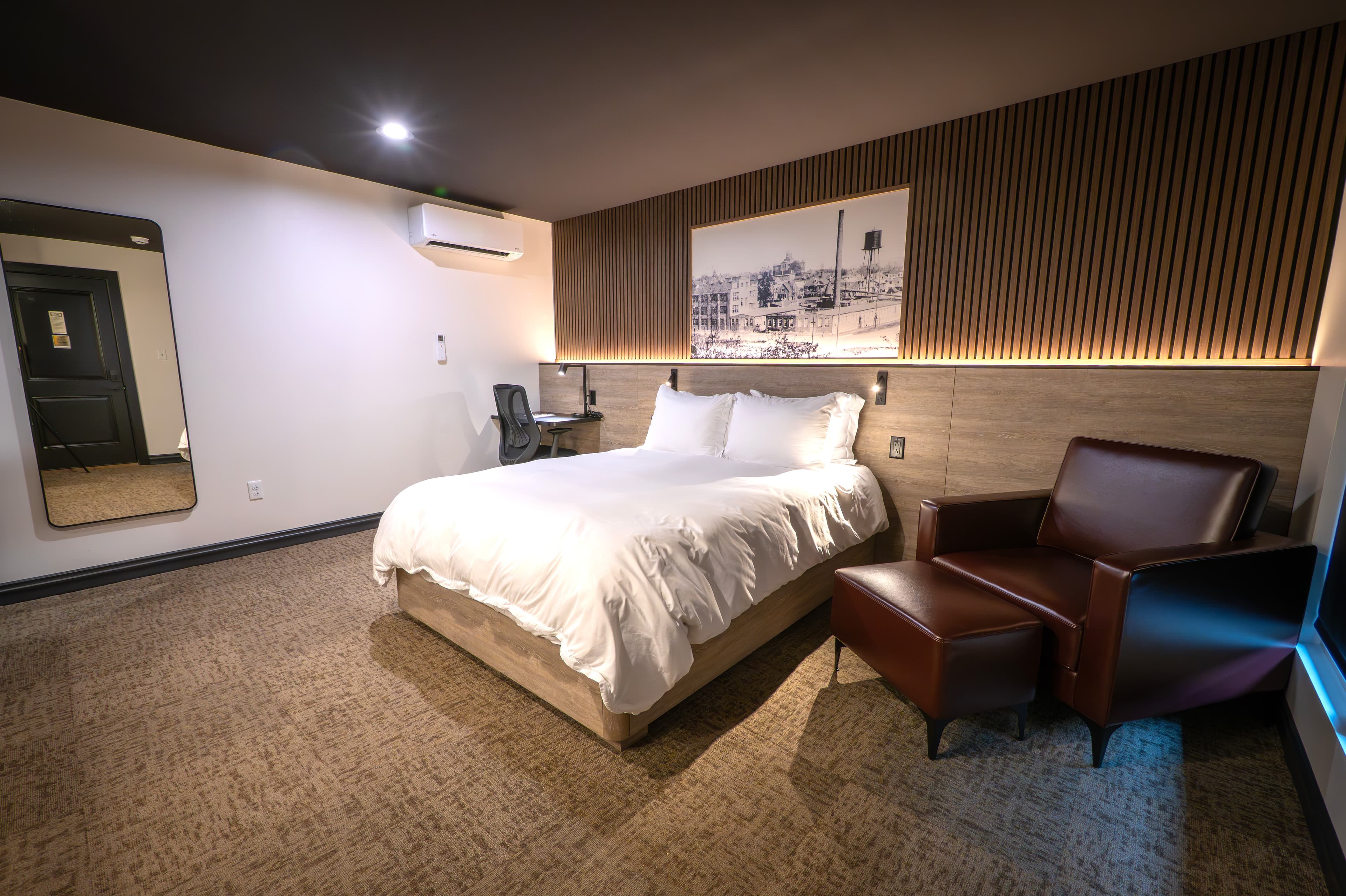 A modern hotel room featuring a queen bed, a armchair, a work desk, and a decorative wall with artwork.