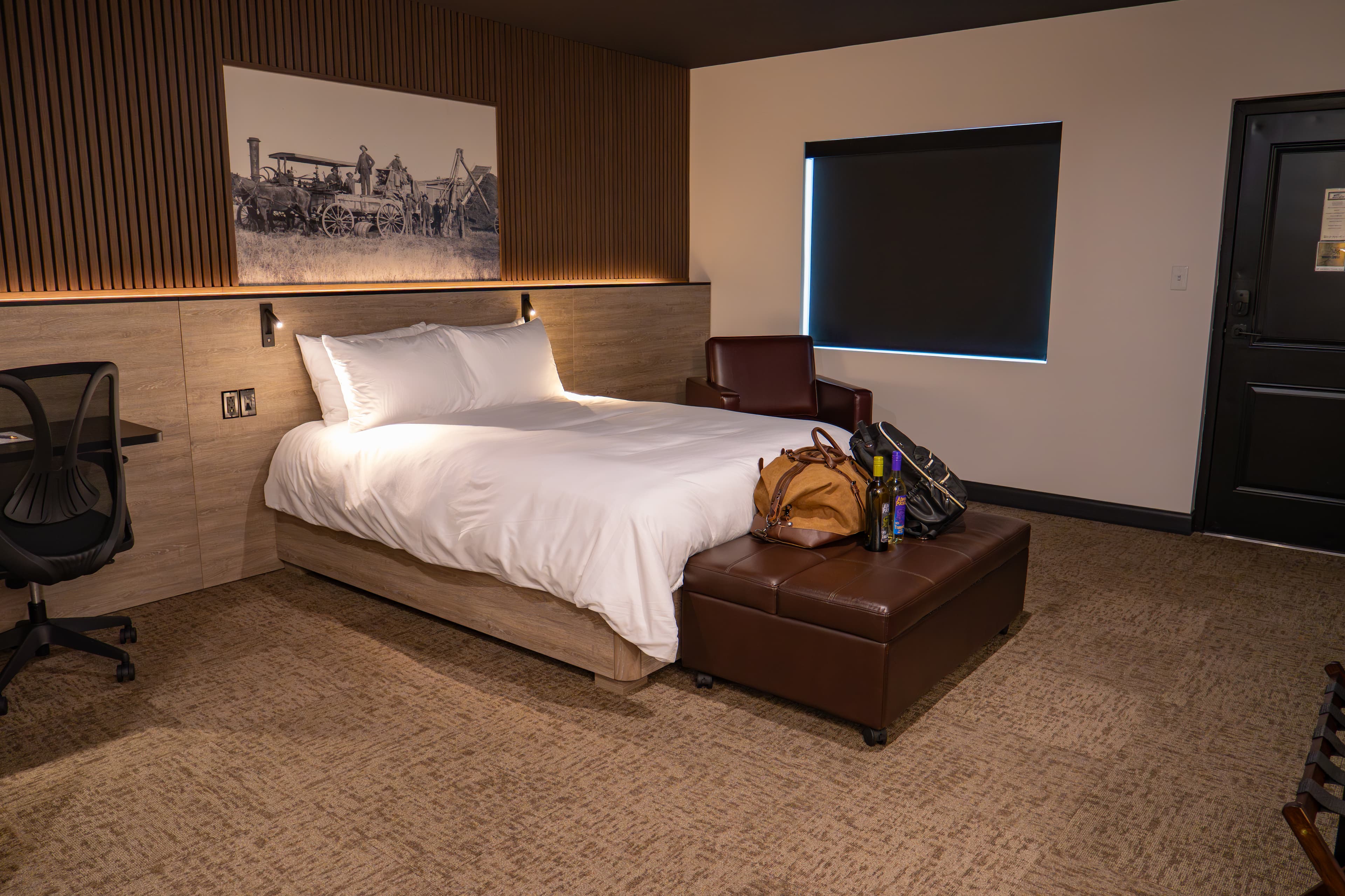 A modern hotel room featuring a queen bed, chair, desk, and a window with shades.