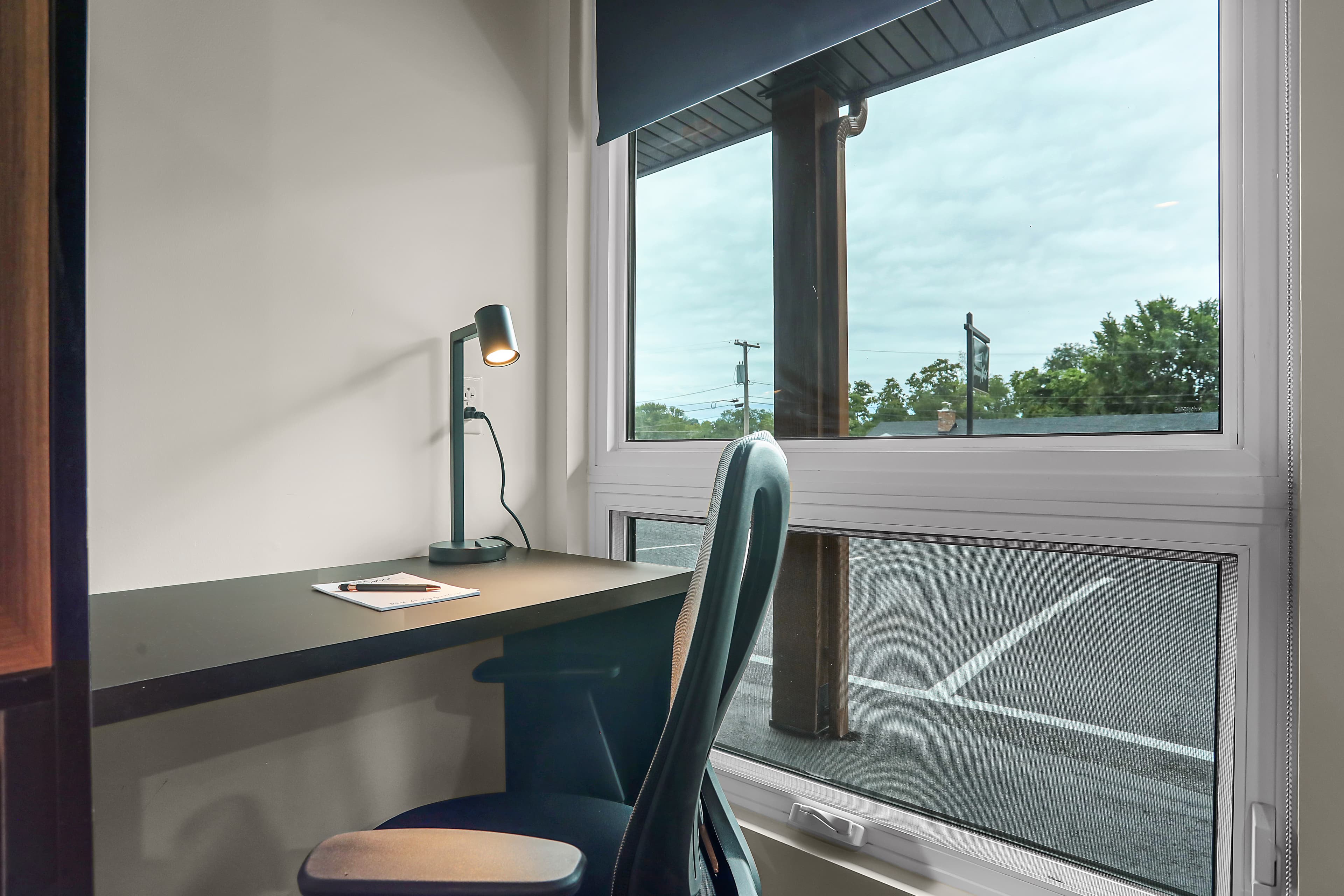 A minimalist desk with a lamp and notebook beside a large window overlooking a cloudy sky and pavement.