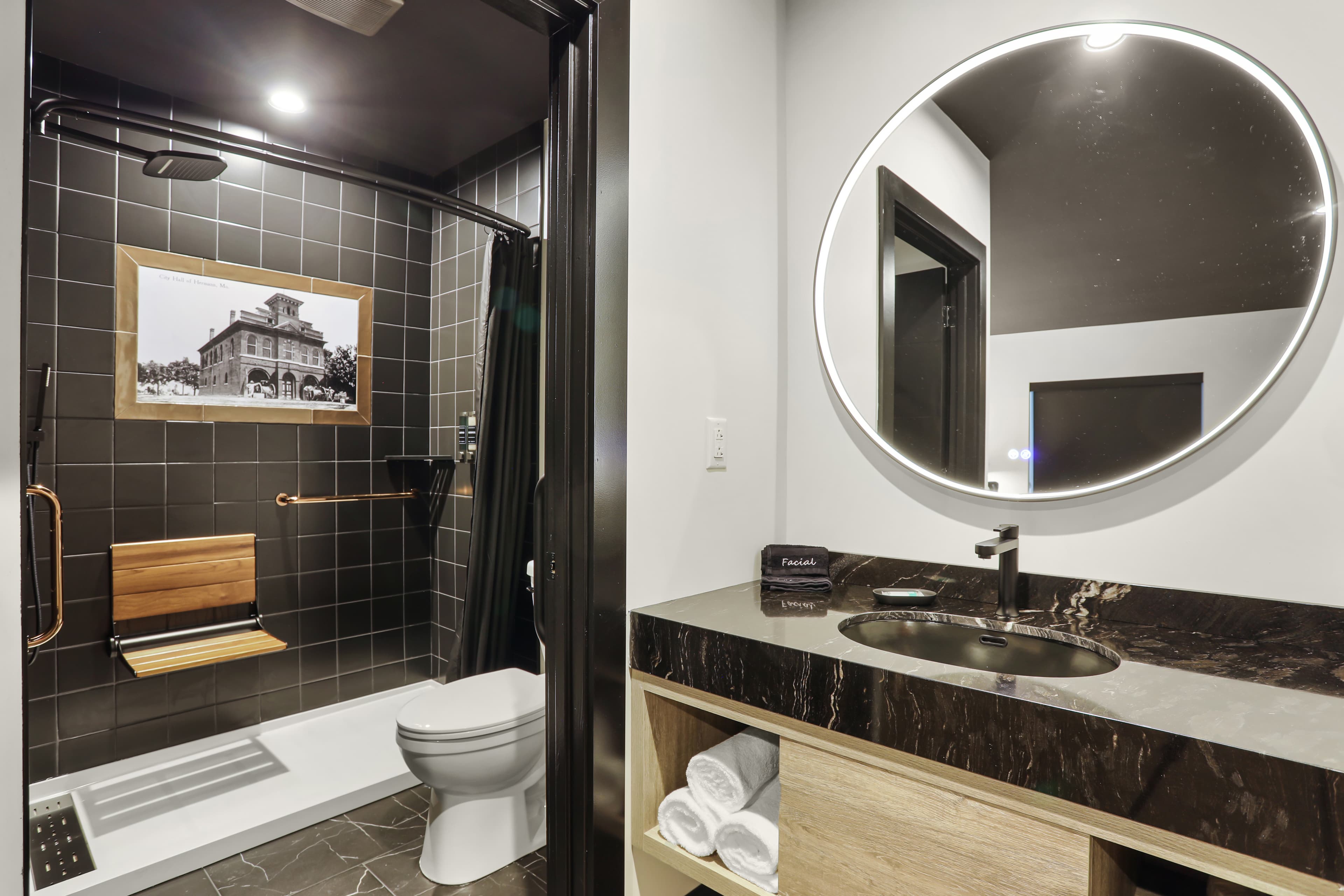A modern bathroom featuring a shower area, wooden bench, round mirror, and a dark marble countertop.