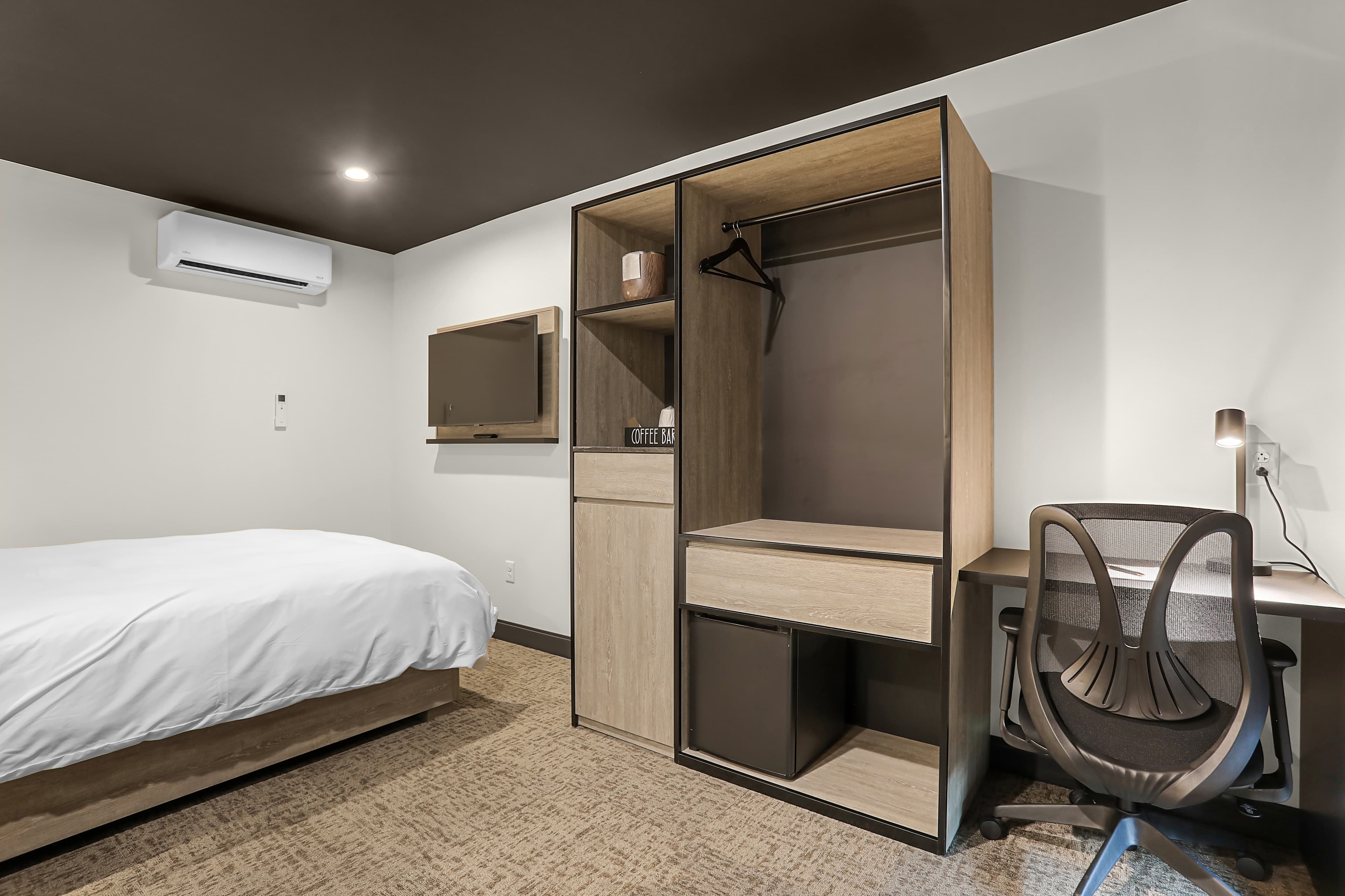 A minimalist hotel room featuring a bed, an entertainment unit, a closet, and a workspace with a chair.