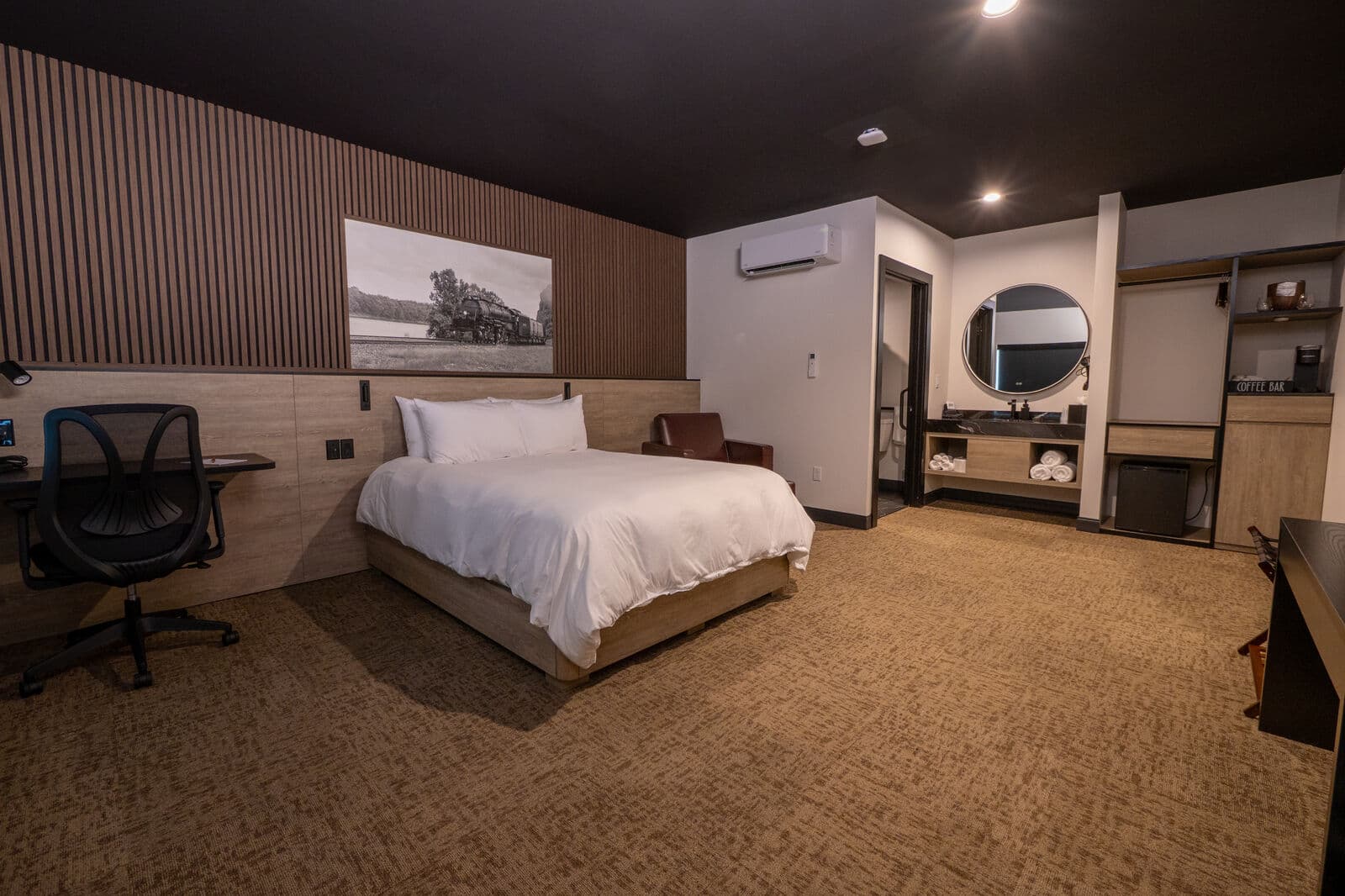 A modern hotel room featuring a bed, desk, and a circular mirror against a neutral color palette.