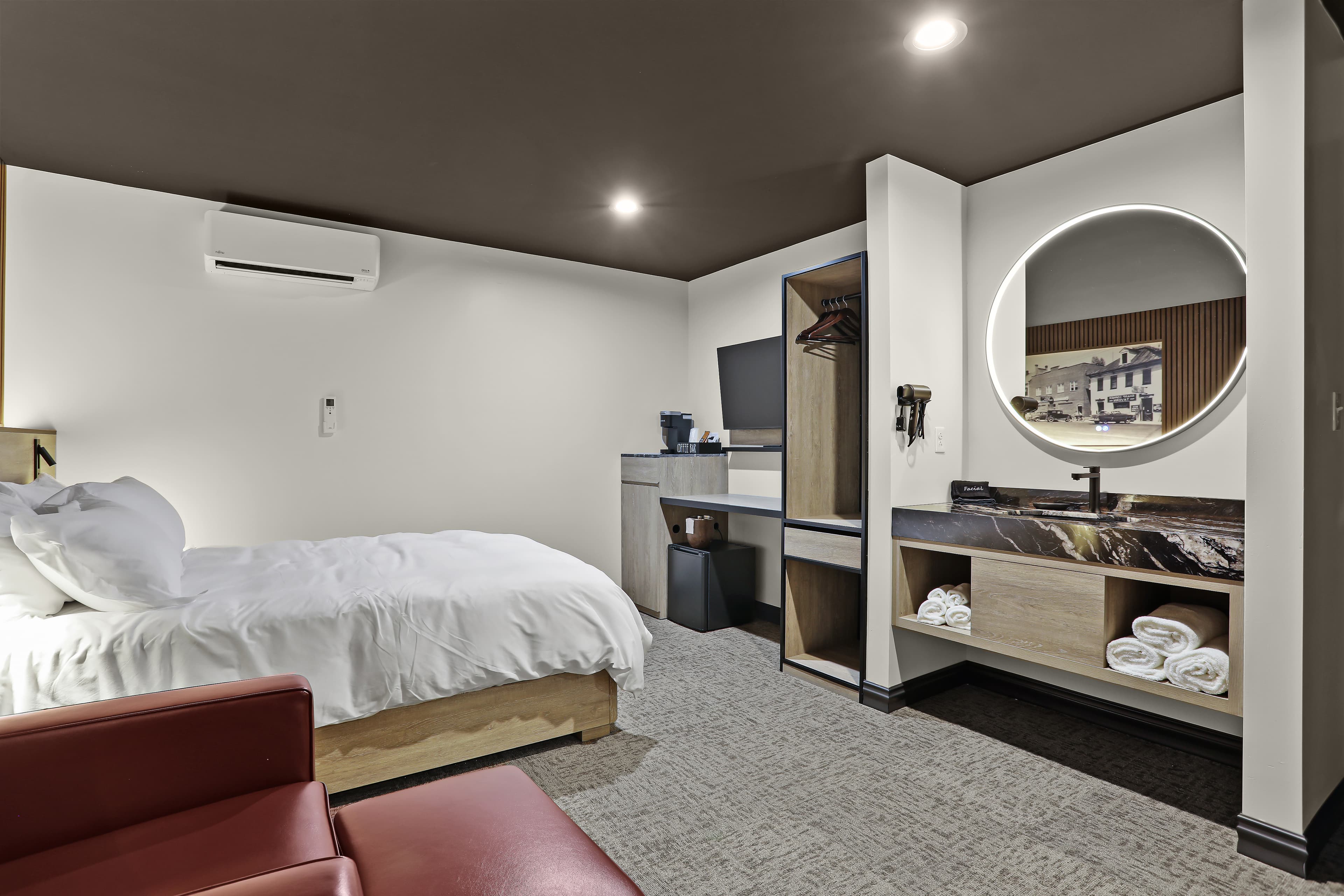 A modern hotel room featuring a double bed, desk, and a round mirror above a sink area.