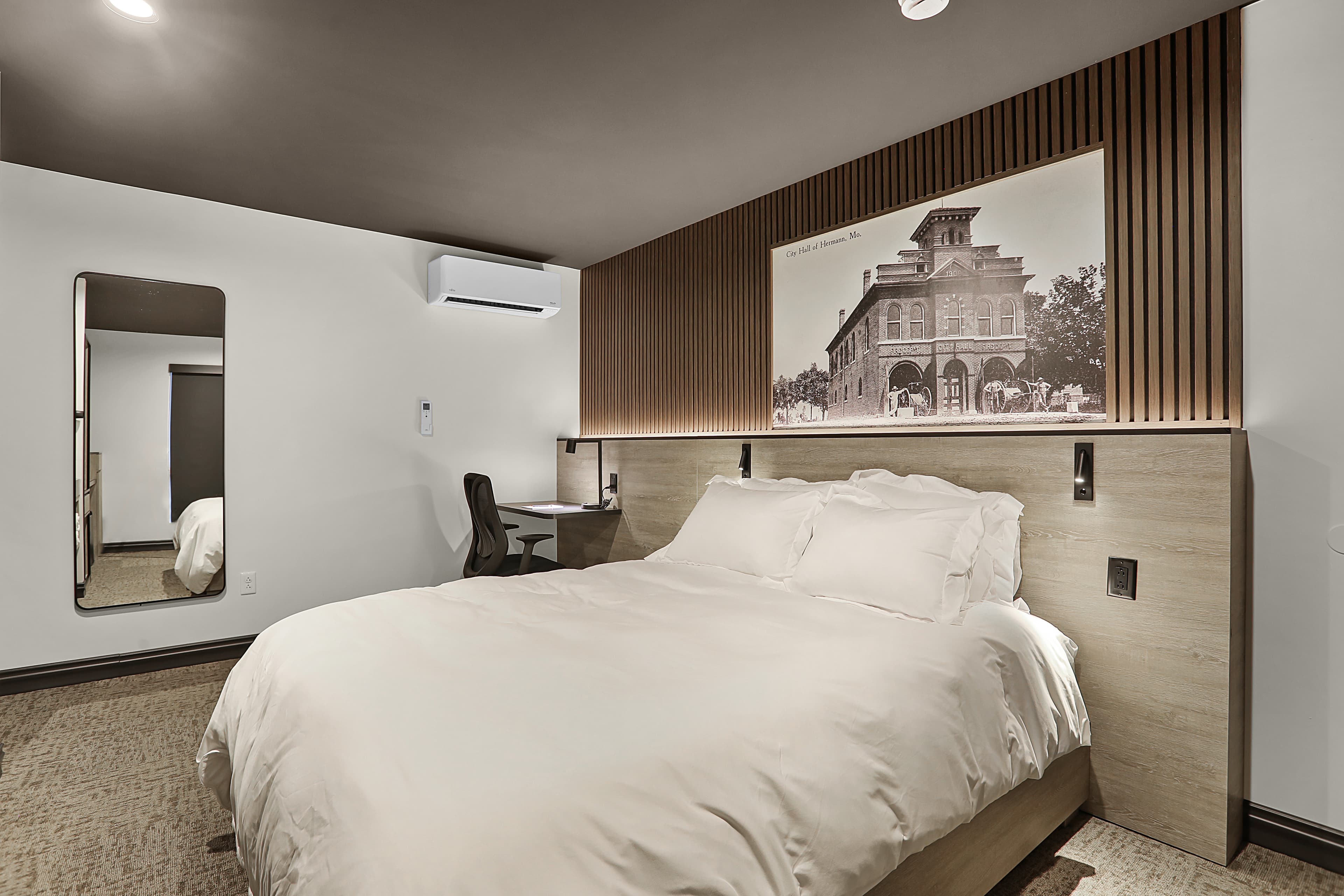 A modern hotel room featuring a queen-sized bed, a mirror, a desk, and a vintage wall art decor.