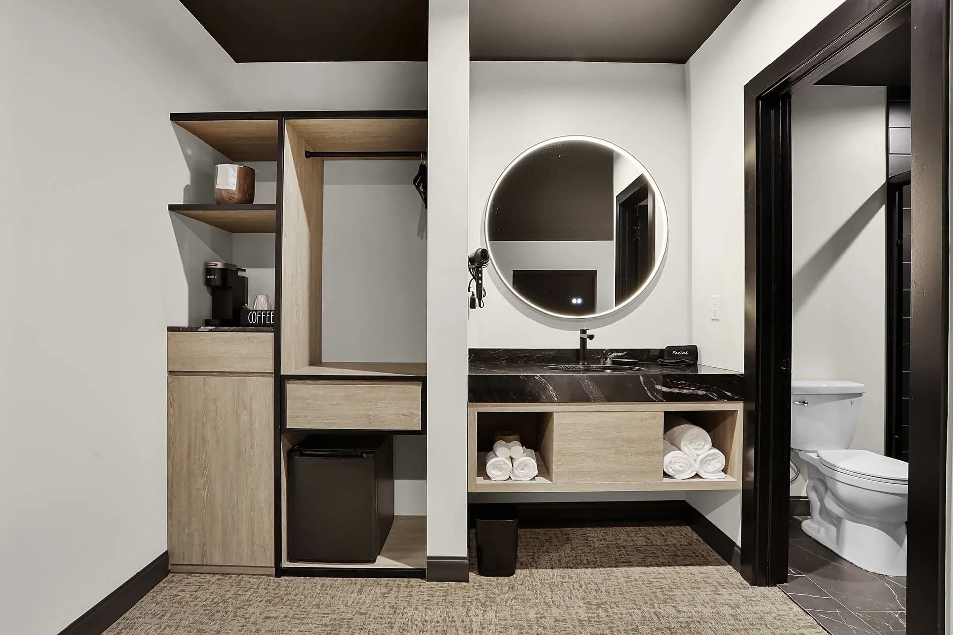A modern bathroom featuring a round mirror, dark countertop, wooden shelving, and a toilet in the background.