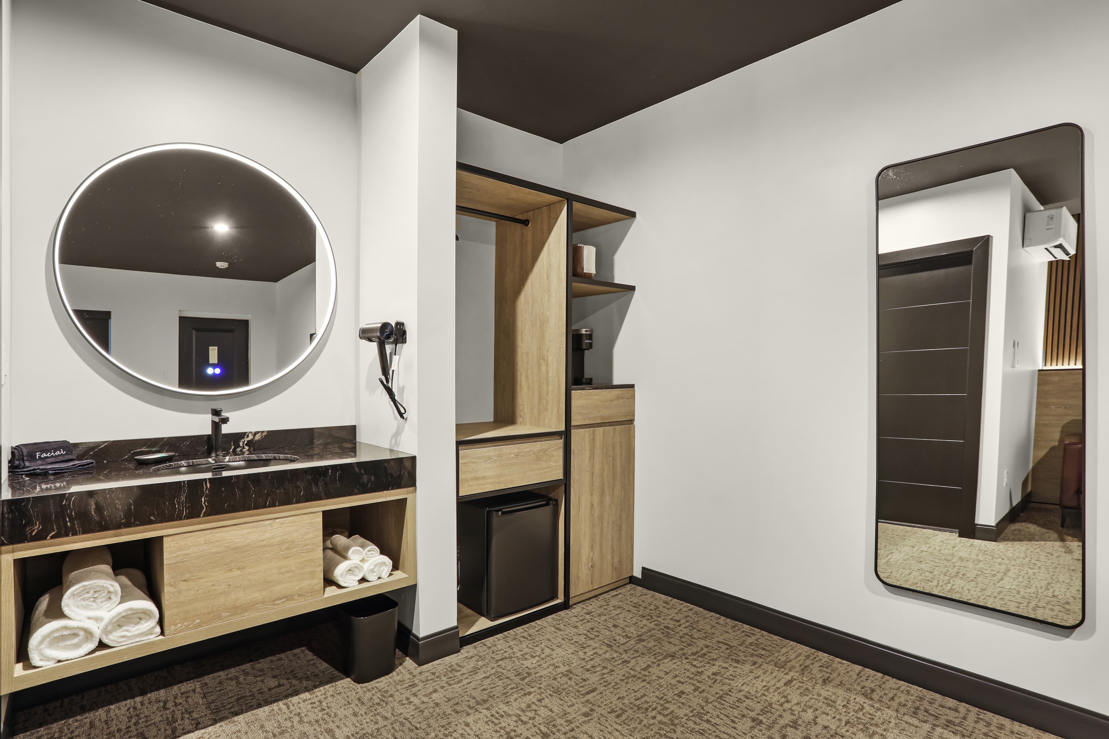 A modern hotel room featuring a circular mirror, dark countertops, wooden cabinetry, and a full-length mirror.