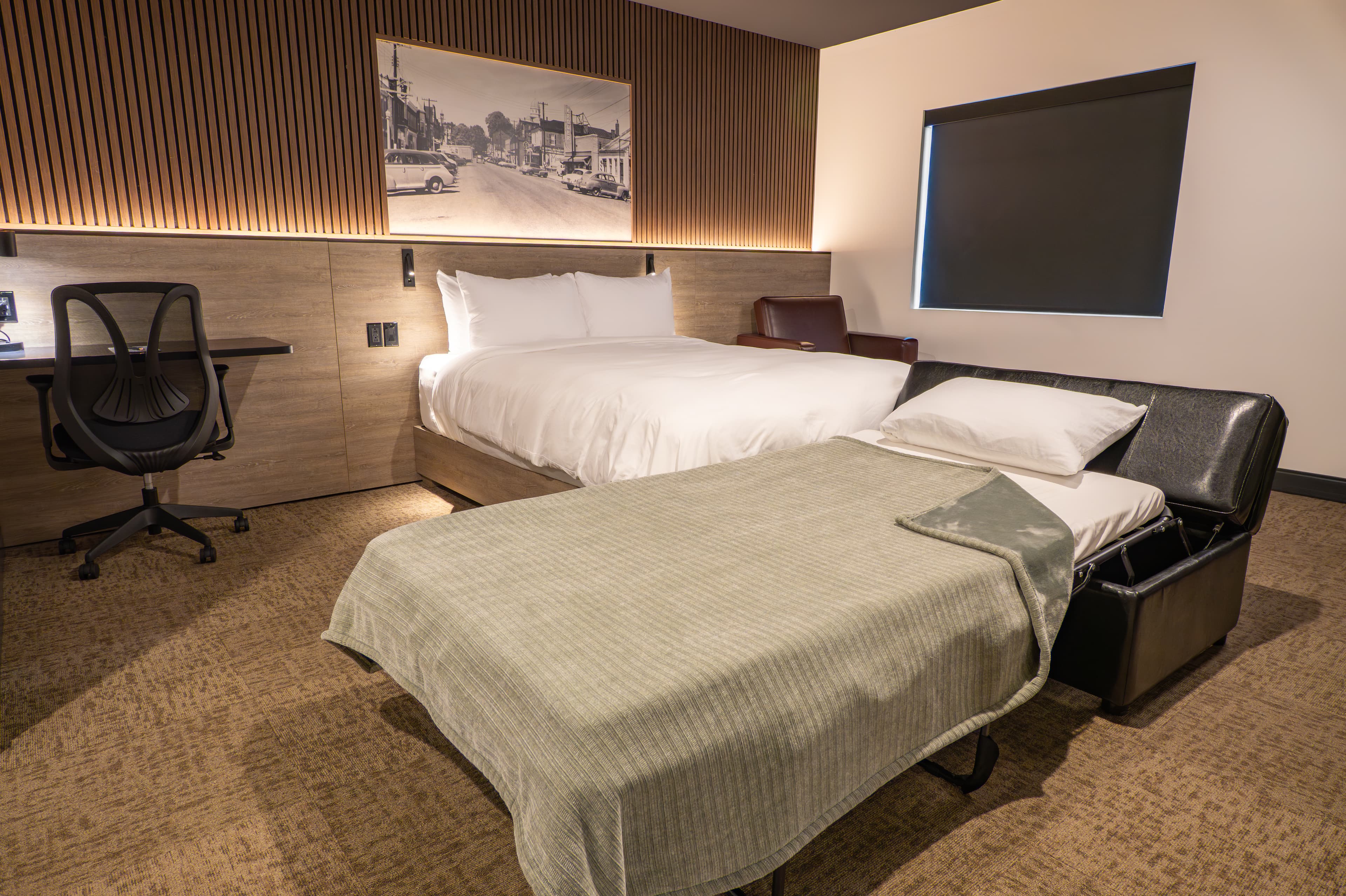 A modern hotel room with a queen bed, a pull-out sofa bed, a desk, and a large window.