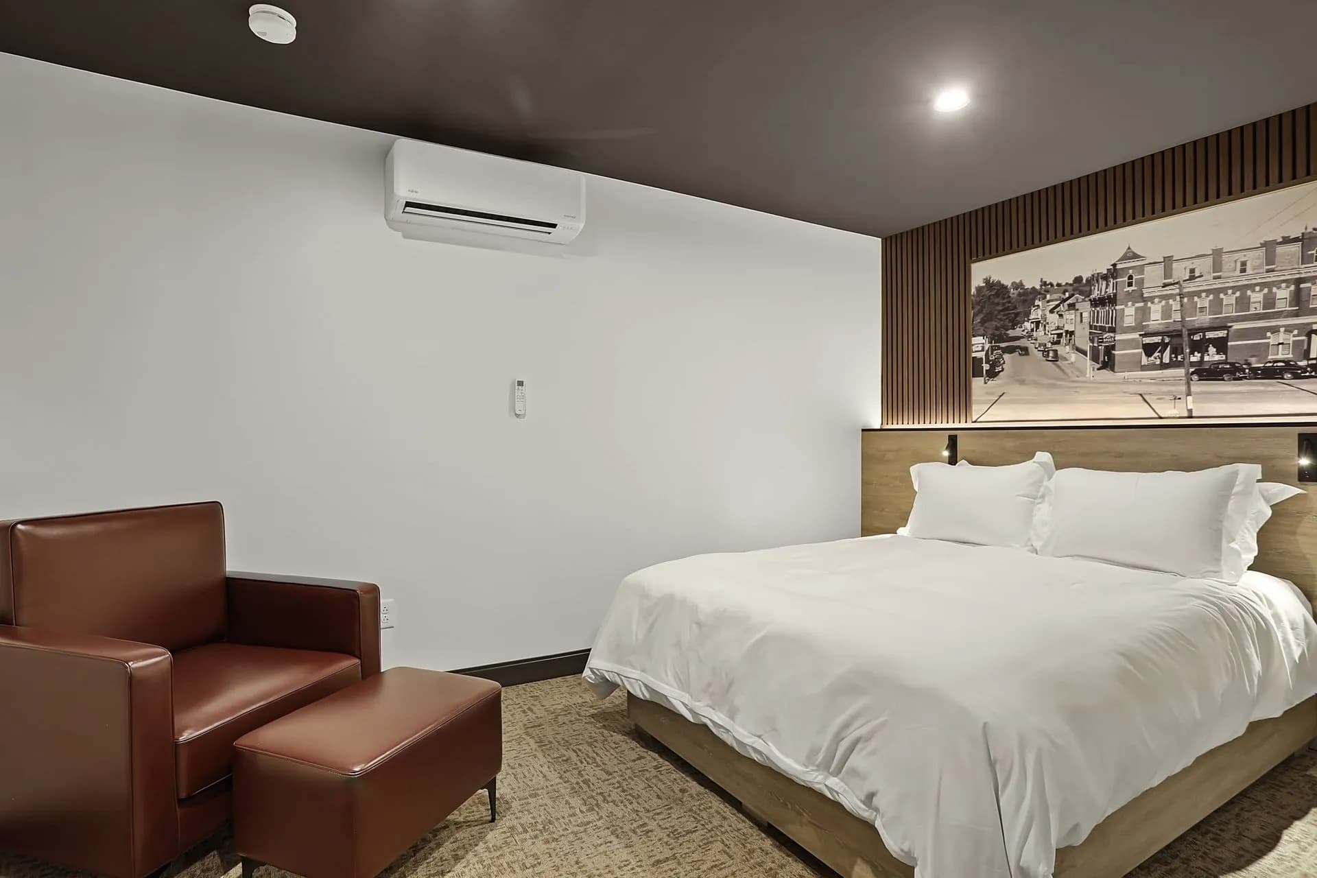 A modern hotel room featuring a queen bed, a red armchair, and a wall-mounted air conditioner.