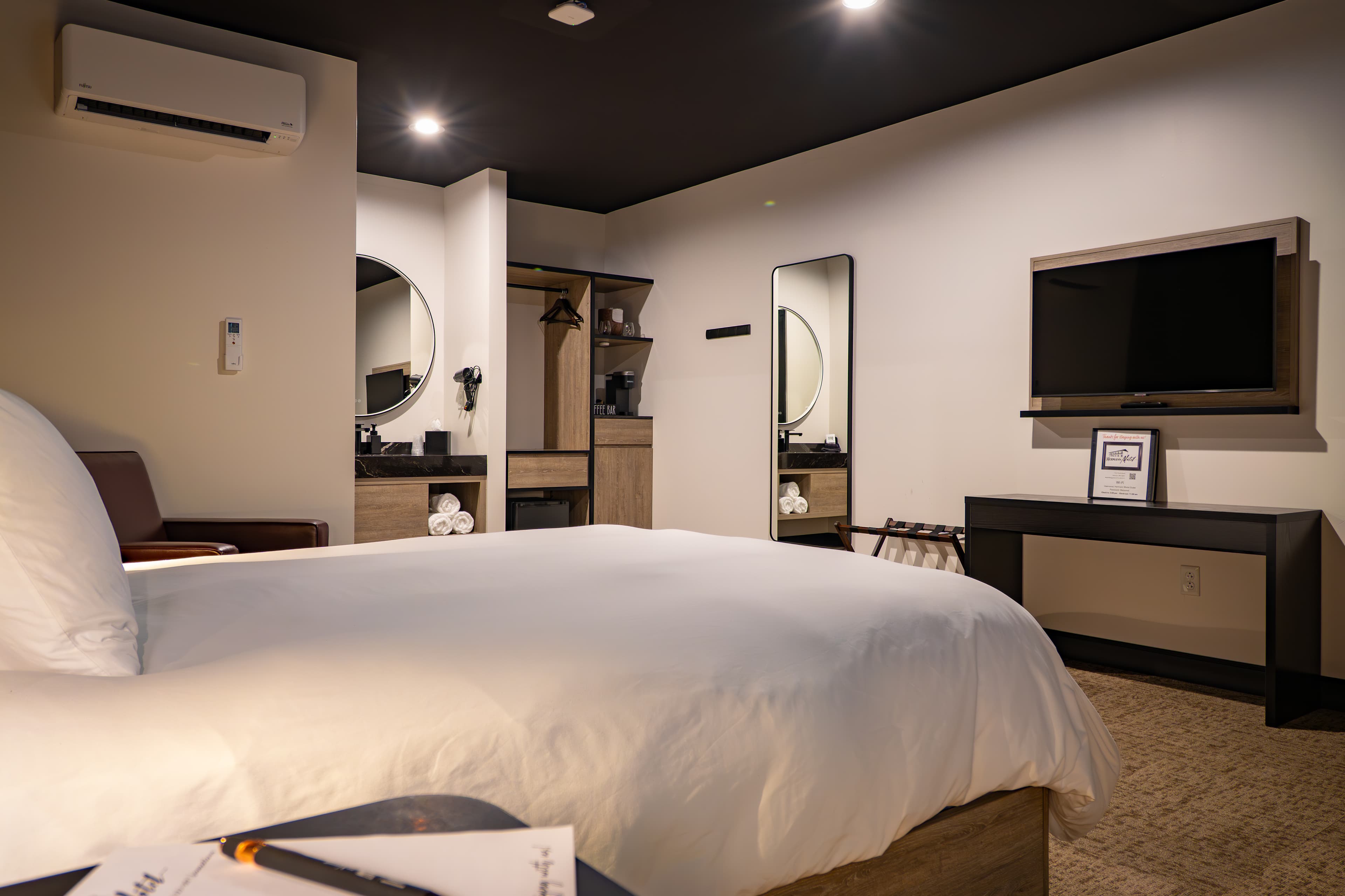 A modern hotel room featuring a bed, a TV, and a mirrored dresser in neutral tones.