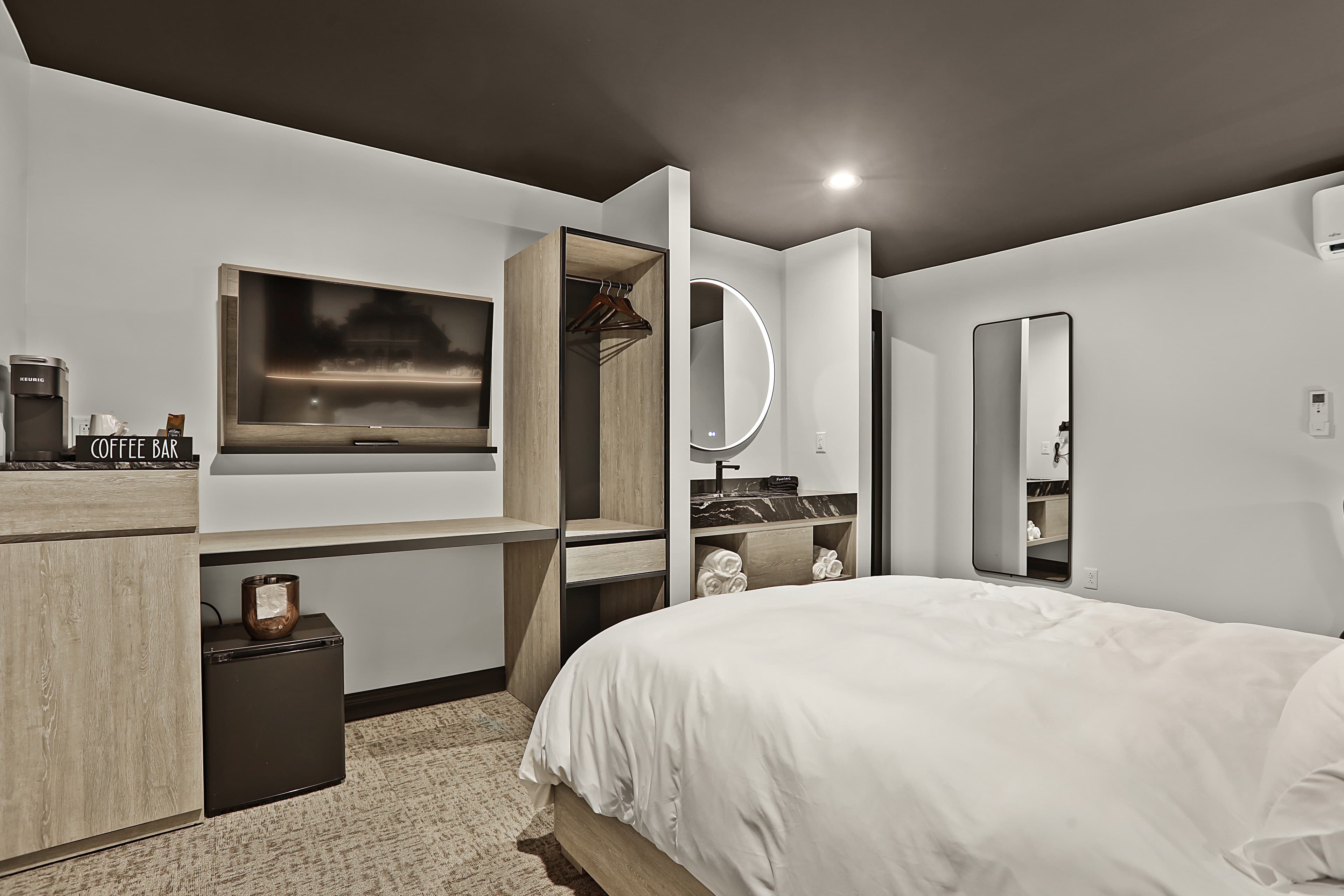 A modern hotel room featuring a bed, TV, mirror, and coffee bar with minimalist decor.